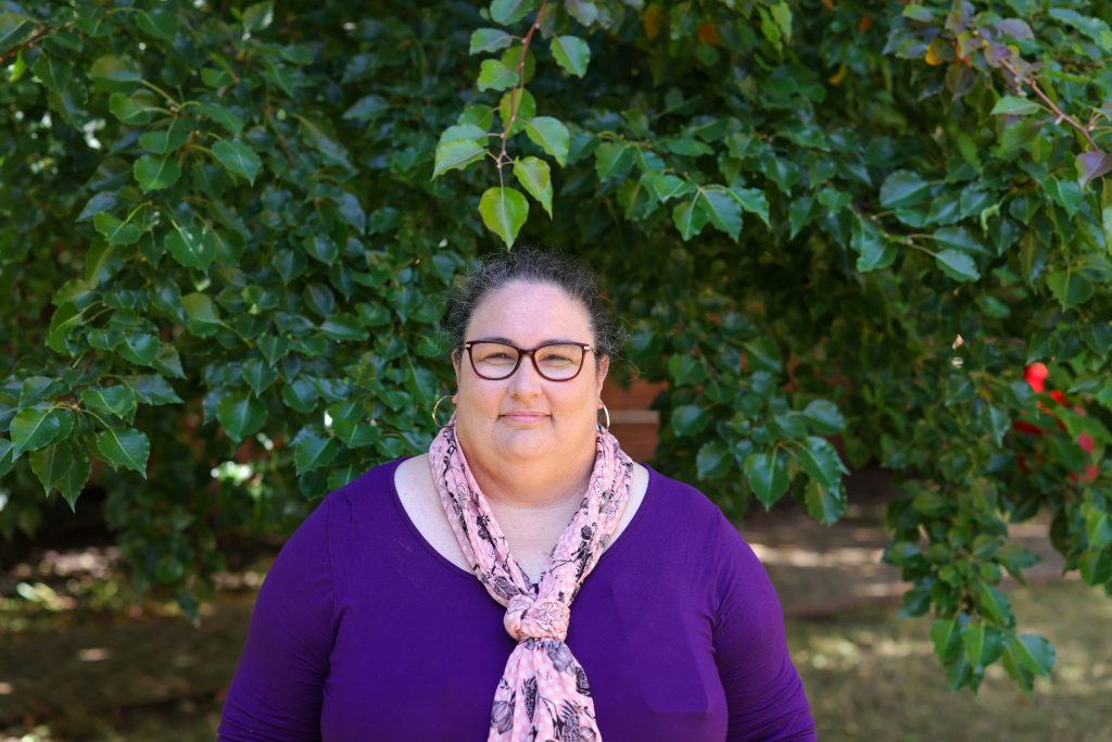 Picture of a younger looking than her age lady in purple, D, standing in front of a green bush
